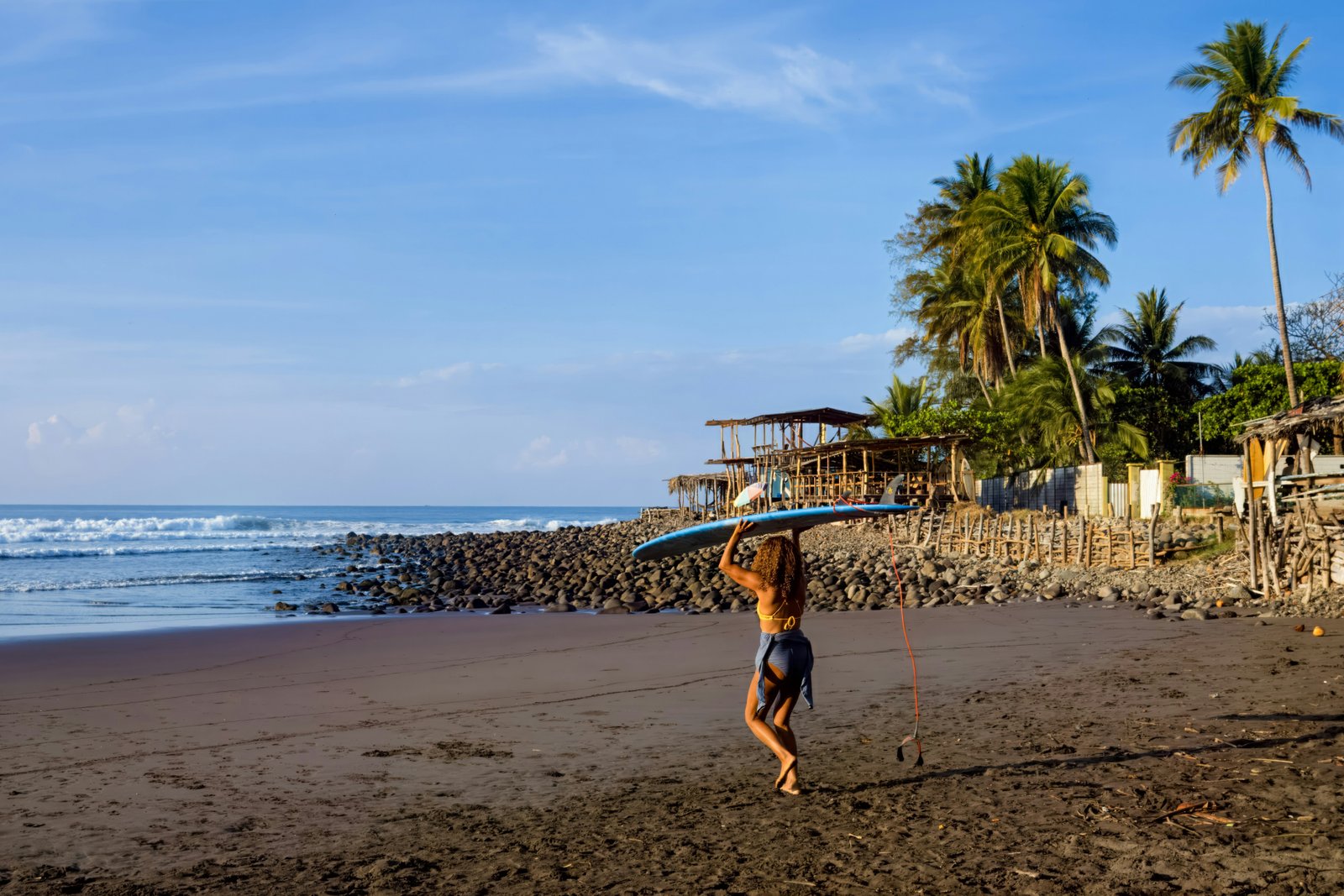 Surfer carrying a board along a volcanic black sand beach, El Salvador