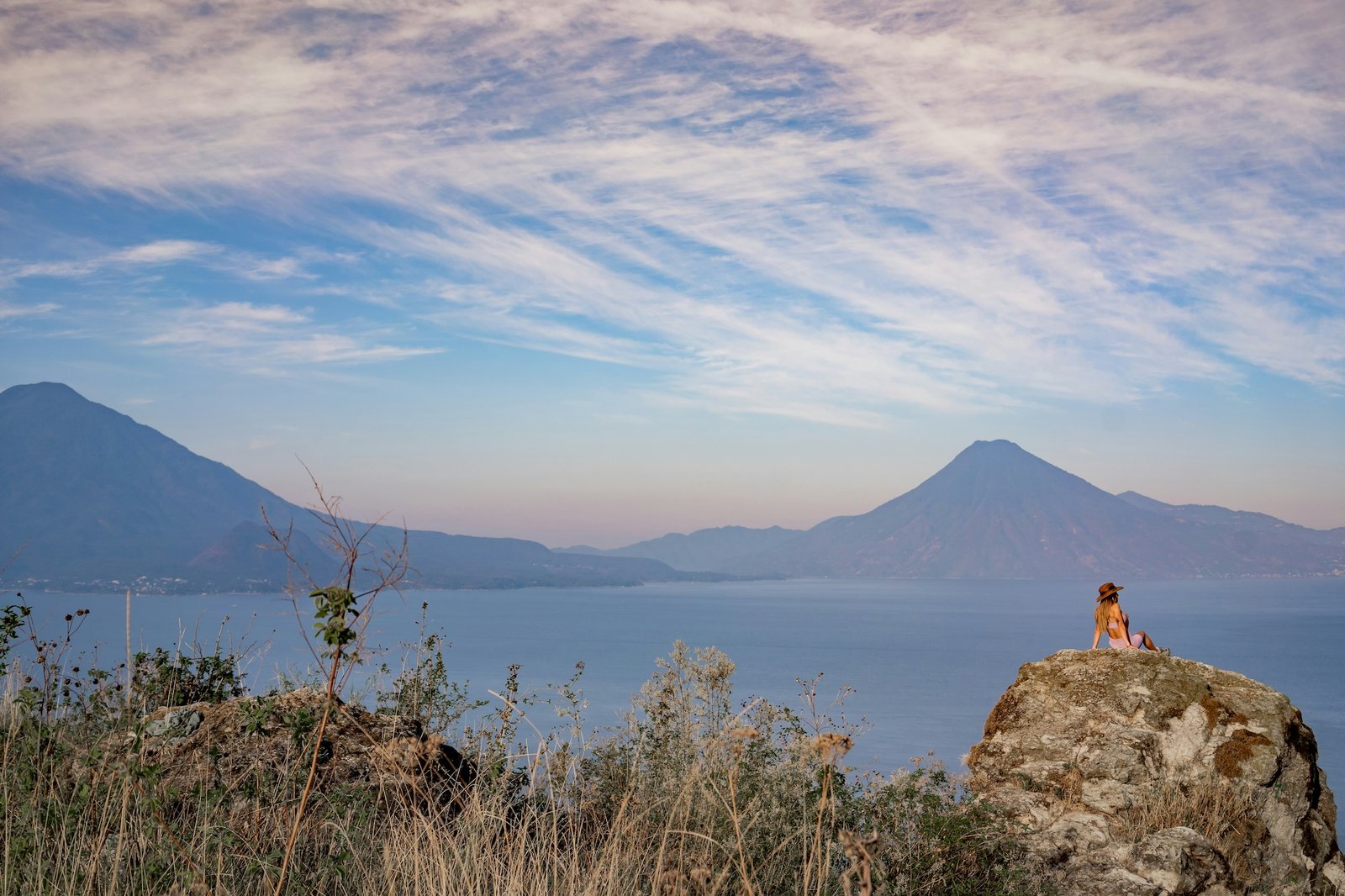 A traveller sits on a rock overlooking Lake Atitlán, two volcanoes rising above the water at dusk
